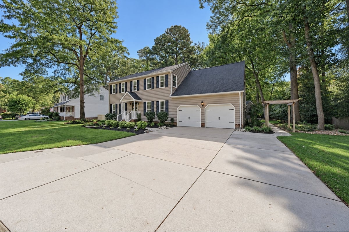 Two-story colonial house with tan siding, dark roof, and two-car garage on a spacious driveway surrounded by large trees.