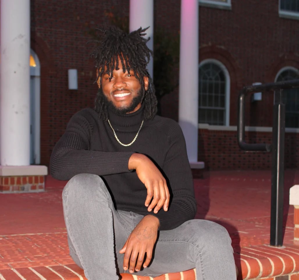 Young man with dreadlocks wearing black sweater and gray jeans, smiling while posing against brick building with white columns